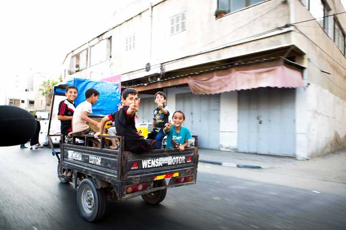 A group of young boys in the back of a small van waving as they ride down a Gazan street.