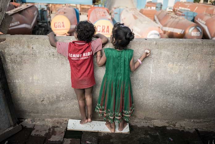 Two young children peering over a wall into a lorry park. Chennai, India — August 2017.