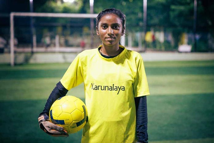 A young Indian woman poses wearing her goal keepers shirt and holding a football.