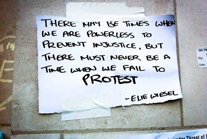 A piece of paper stuck the the pavement on a demo reads, "There may be times when we are powerless to prevent injustice, but there must never be a time when we fail to protest."