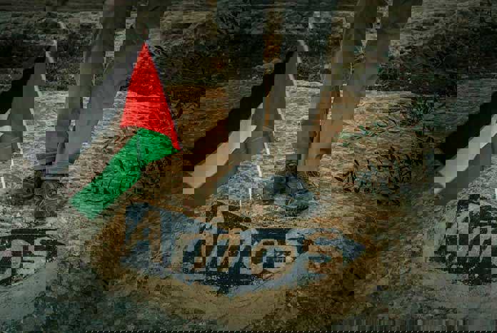 A Palestinian flag planted in the sand near a rock that has the word 'Amos' written on it.