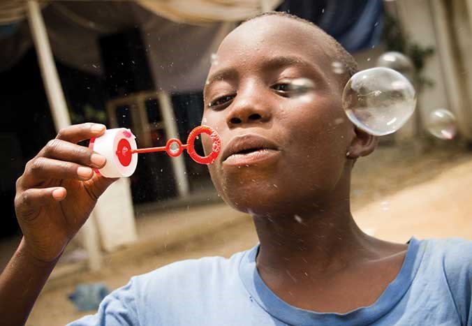 A young boy in Burundi blows bubbles in the air while smiling.