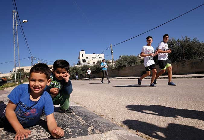 Two young boys sitting by the side of a road and laughing in Bethlehem, Palestine, as two male marathon runners run by smiling.