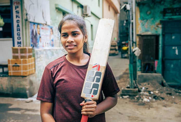 A young Indian woman holds a cricket bat over her shoulder.